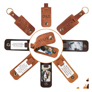 May include: A collection of brown leather keychains featuring metal snap closures and metal rings. The leather is engraved with text including DAD, PAPA, and DADDY. Some keychains contain rectangular photo inserts or white cards with printed messages.