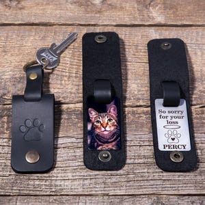 May include: Three black leather keychains with metal snaps. One features a debossed paw print and a silver key. Another displays a color portrait of a tabby cat. The third shows a silver tag with the text So sorry for your loss, a halo, a paw print, and the name PERCY.