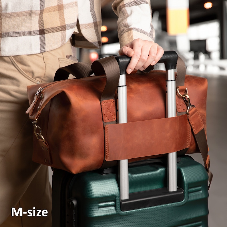 May include: A brown leather holdall with dark brown nylon handles and a matching leather luggage strap, secured over the silver telescopic handle of a dark green hard-shell suitcase. The text M-size is visible in the bottom left corner.