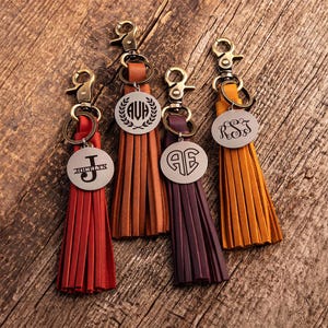 May include: Four leather tassel keychains with antique brass lobster clasps and circular metal tags. The tags feature engraved monograms and names, including JOCELYN, AVH, AE, and RSJ. The tassels are red, tan, dark purple, and mustard yellow.