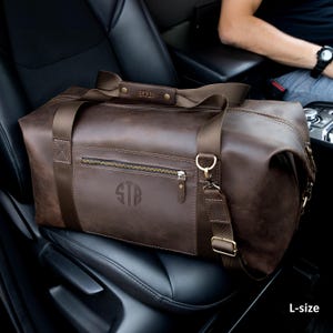 May include: A dark brown leather duffel bag featuring a horizontal zippered front pocket, a monogrammed circular logo with the letters STB, and a matching leather handle wrap embossed with STB. The bag includes bronze-toned hardware and straps.