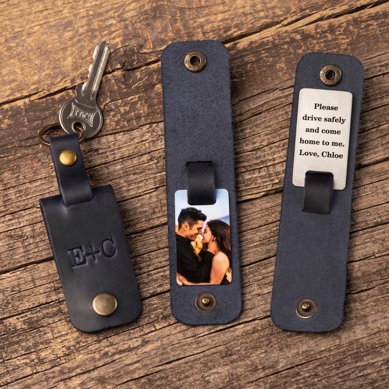 May include: Three dark blue leather keychains with brass hardware. One features the embossed letters E+C, one displays a color photograph of a couple, and one shows a metal tag engraved with the text: Please drive safely and come home to me. Love, Chloe.