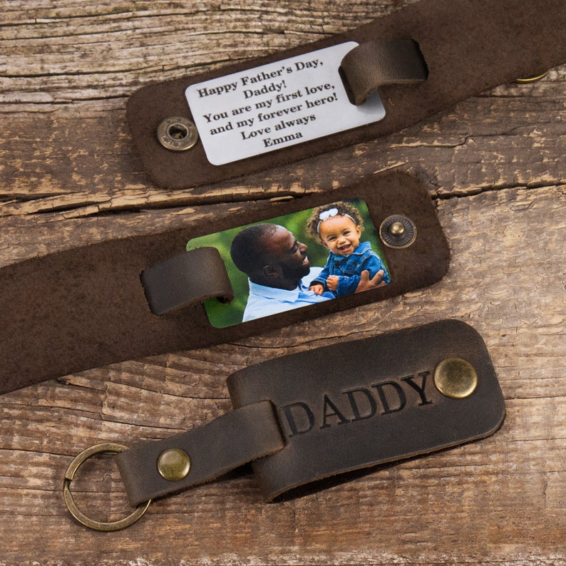 May include: Three dark brown leather accessories with brass-toned metal snaps. One features a silver metal plate with the text: Happy Father's Day, Daddy! You are my first love, and my forever hero! Love always Emma. Another shows a color photo of a person holding a child. The third is a keychain embossed with the word DADDY.