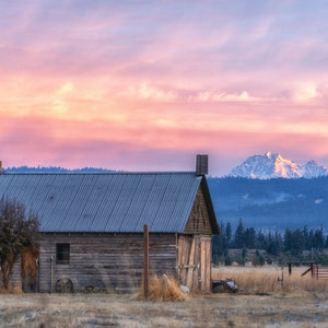 May include: A weathered wooden barn with a metal roof sits in a field with a snow-capped mountain range in the background. The sky is a vibrant pink and purple.