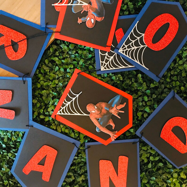 May include: A red and blue banner with the word "AND" in red glitter letters. Each letter is on a separate banner with a black background and a blue border. The banners are decorated with a white spider web design and a red spider-man character.