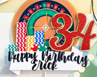 May include: A white cake topper with a green and gold roulette wheel, red and gold number 34, and the text "Happy Birthday Erick".