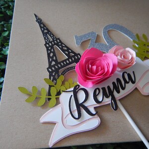 May include: A cake topper featuring a black silhouette of the Eiffel Tower, two pink paper flowers, green leaves, and a white banner with the name "Reyna" in black script.