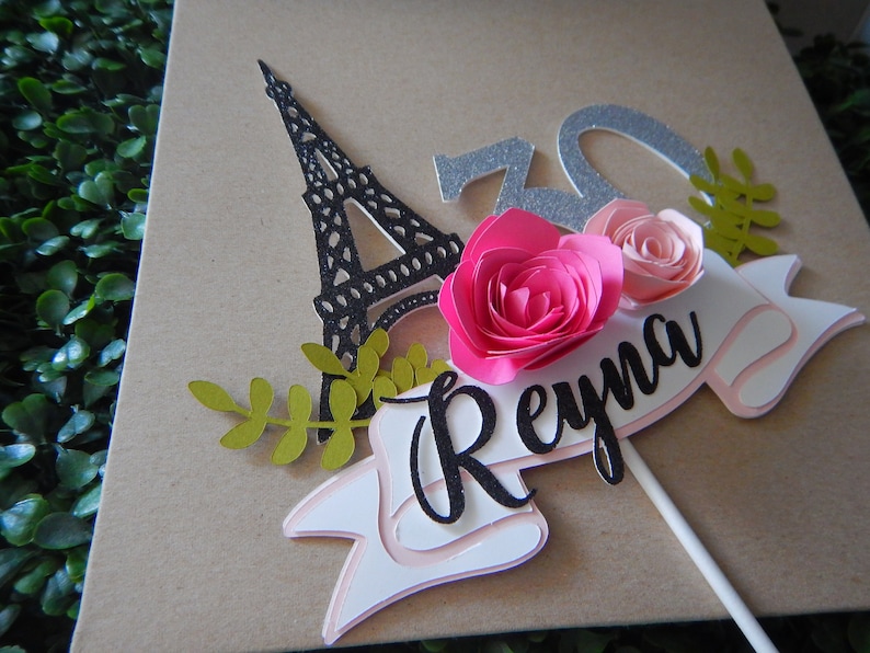May include: A cake topper featuring a black silhouette of the Eiffel Tower, two pink paper flowers, green leaves, and a white banner with the name "Reyna" in black script.