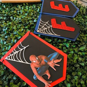 May include: A black and red Spiderman banner with a silver spider web design. The banner is made of cardstock and has a red border. The banner is perfect for a Spiderman themed party.