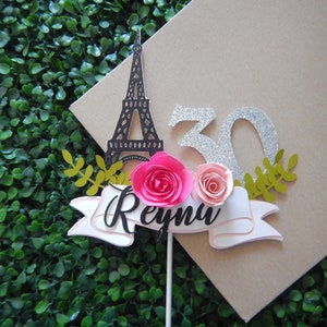 May include: A cake topper featuring a black silhouette of the Eiffel Tower, a silver glitter 30, two pink paper flowers, and a white banner with the name "Reyna" in black script.