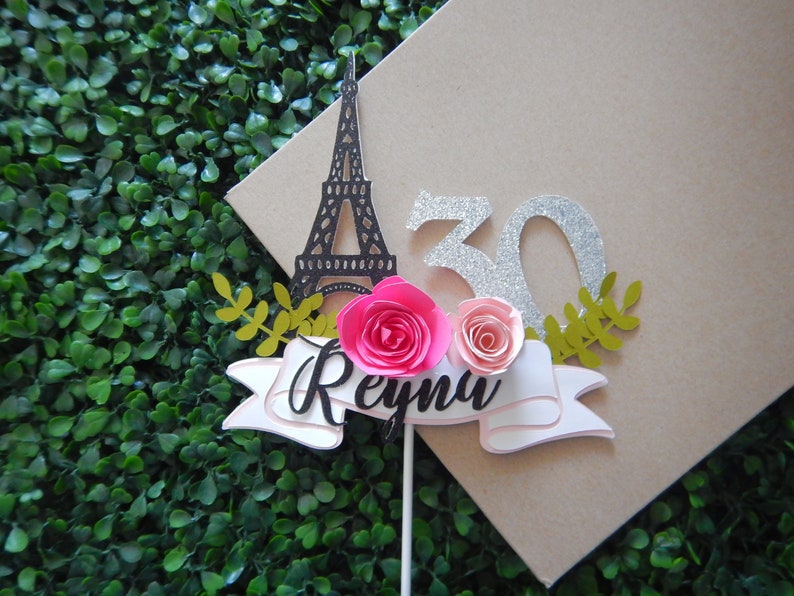 May include: A cake topper featuring a black silhouette of the Eiffel Tower, a silver glitter 30, two pink paper flowers, and a white banner with the name "Reyna" in black script.