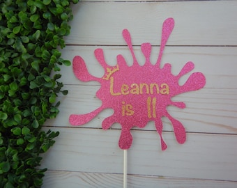 May include: Pink glitter paint splatter cake topper with a gold crown and the text "Leanna is 11".