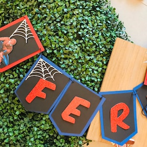 May include: A red and blue banner with the word "FER" in red glitter letters. Each letter is on a separate black card with a white spiderweb design. The banner is decorated with a red and black Spiderman character.