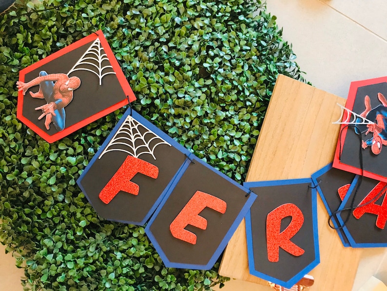 May include: A red and blue banner with the word "FER" in red glitter letters. Each letter is on a separate black card with a white spiderweb design. The banner is decorated with a red and black Spiderman character.