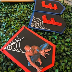 May include: A black and red Spiderman banner with a silver spider web design. The banner is made of cardstock and has a red border. The banner is perfect for a Spiderman themed party.