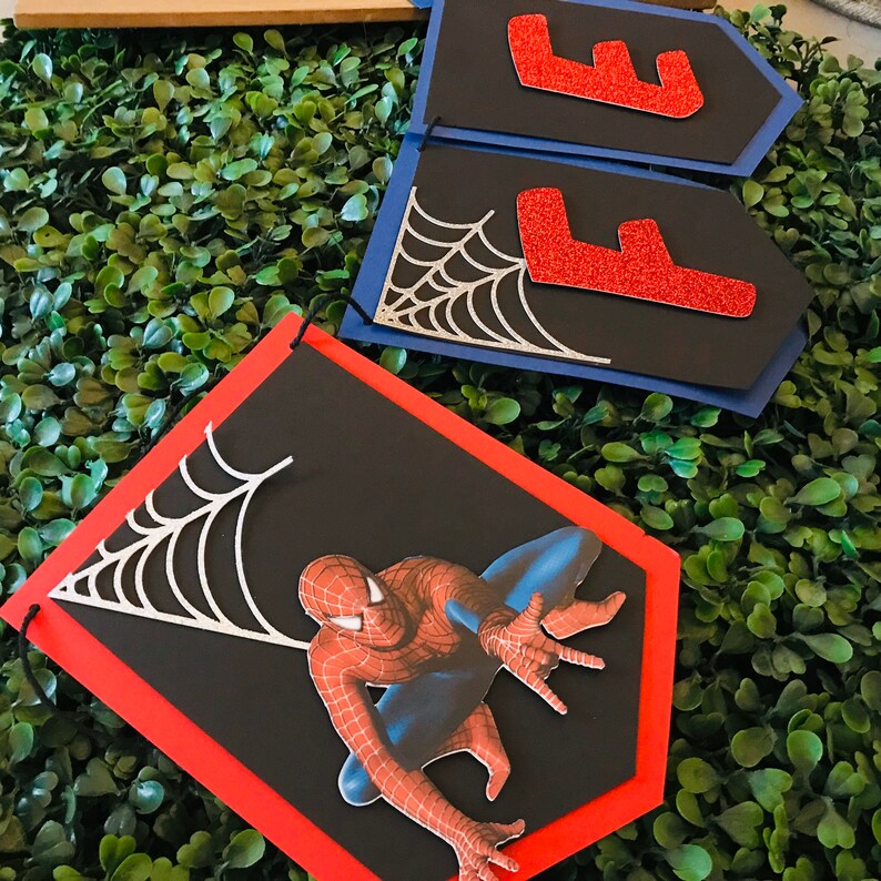 May include: A black and red Spiderman banner with a silver spider web design. The banner is made of cardstock and has a red border. The banner is perfect for a Spiderman themed party.