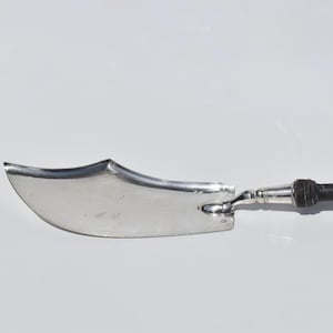 May include: A silver cheese knife with a curved blade and a dark wooden handle. The handle is connected to the blade with a silver metal piece. The knife is designed for cutting cheese and other soft foods.