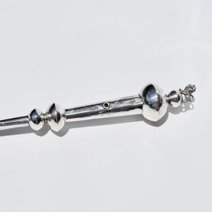 Może przedstawiać: Srebrzysty, wydłużony przedmiot o dekoracyjnym wzorze. Posiada szereg sferycznych i cylindrycznych kształtów, zakończonych ozdobnym elementem. Drugi koniec ma konstrukcję przypominającą śmigło. Wymiary w centymetrach.