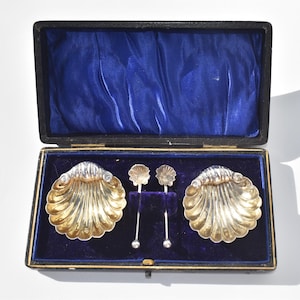 May include: A set of two silver-toned shell-shaped dishes and two matching small spoons with shell-shaped bowls, presented inside a rectangular black storage case lined with deep blue velvet fabric, featuring a hinged lid and gold-toned trim.