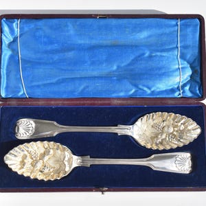 May include: Two silver-toned serving spoons with embossed fruit and leaf patterns on the bowls and shell motifs on the handles. The spoons are presented in a rectangular case with a dark purple exterior and a vibrant blue velvet interior lining.