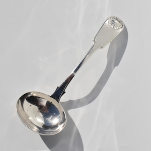May include: A polished silver ladle featuring a round bowl and a long, slender handle. The handle terminates in a flat, rectangular end decorated with a raised, embossed shell motif at the top. The metal surface is reflective and smooth.