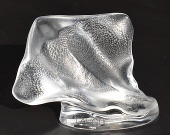 May include: A clear glass sculpture with a textured, scale-like pattern. The base is a clear, curved shape with the text "LALIQUE PARIS" etched on it. The sculpture has a flowing, organic form.
