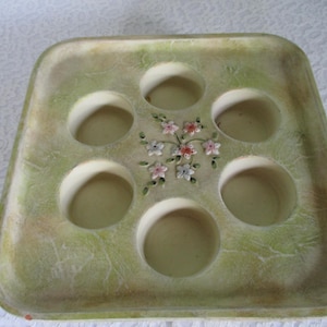 May include: A vintage, square-shaped egg tray with six circular indentations. The tray has a mottled green and beige finish and is decorated with a floral design in the center. The flowers are pink, white, and red.