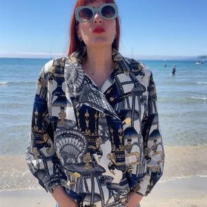 May include: A patterned button-up shirt with a navy blue, white, and gold design. The shirt features a unique print of ornate architectural elements. The shirt has long sleeves and gold buttons. The person is wearing blue sunglasses and denim shorts.