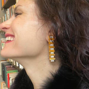 May include: Gold-coloured rectangular drop earrings with clear, rectangular stones. The earrings are worn by a person with dark hair and a black fur-like coat. A bookshelf is visible in the background.
