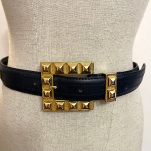 May include: A black leather belt with a large, gold-toned buckle featuring a square design with pyramid-shaped studs.