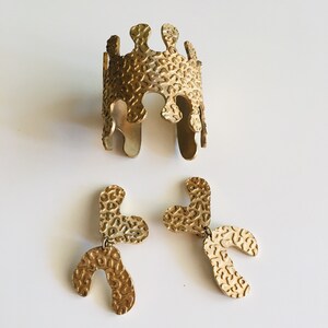 May include: A set of gold-toned jewelry featuring a textured cuff bracelet and a pair of dangling earrings. The bracelet and earrings have a unique, organic shape with a textured surface.