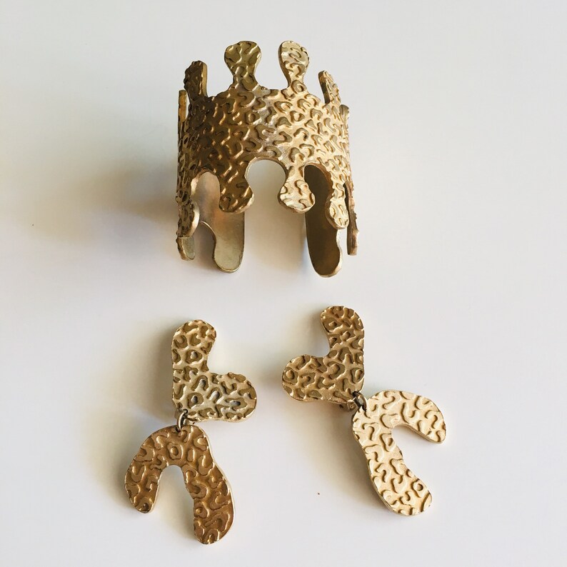 May include: A set of gold-toned jewelry featuring a textured cuff bracelet and a pair of dangling earrings. The bracelet and earrings have a unique, organic shape with a textured surface.