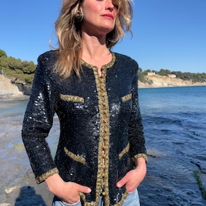 May include: A black sequined jacket with gold detailing, including trim and buttons. The jacket is worn with blue jeans. The gold trim outlines the collar, pockets, and button closures. The background shows a blue sky and the sea.