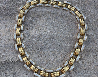 May include: A gold-toned necklace with a linked design, featuring alternating sections of solid gold and sections encrusted with small, clear gemstones. The necklace is displayed on a sandy surface, with the sunlight reflecting off the jewelry.