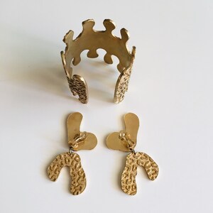 May include: A set of gold-toned jewelry featuring a wide cuff bracelet and a pair of clip-on earrings. The bracelet and earrings have a textured, organic design.