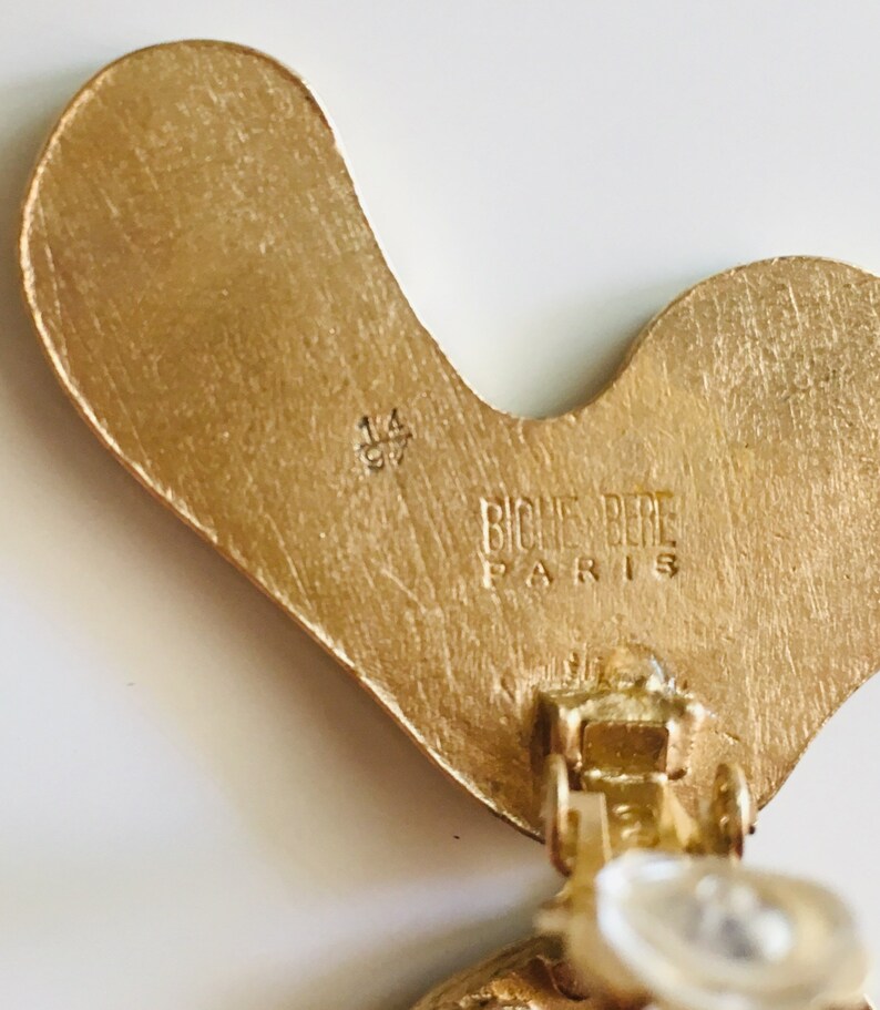 May include: Gold-tone metal earrings with a unique, abstract shape. The back of the earrings feature a clasp closure and the inscription "Biche Pere Paris 14 CT".