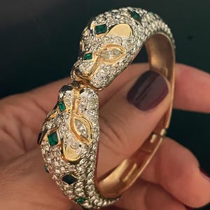 May include: A hinged bangle bracelet featuring two panther heads meeting at the center. The bracelet is crafted from gold-tone metal, fully encrusted with clear pavé crystals, and accented with marquise-cut clear stones and square-cut green stones.