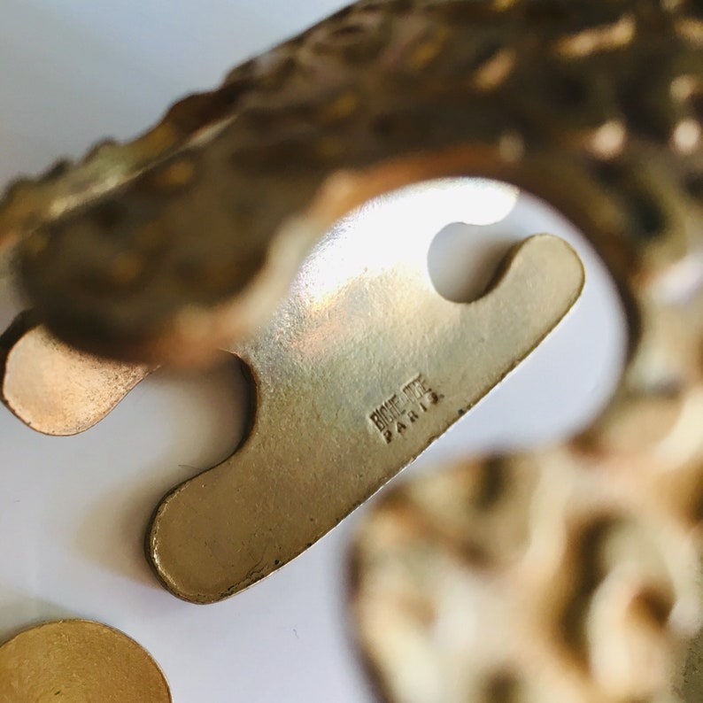 May include: Close-up of a gold-toned metal napkin ring with a detailed design. The ring is marked "Bijouterie Paris" on the underside.