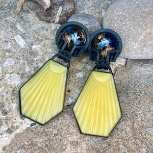 Pode incluir: Um par de brincos geométricos amarelos e pretos com um design de raios de sol. Os brincos têm uma moldura de metal preto e um centro de resina amarela.