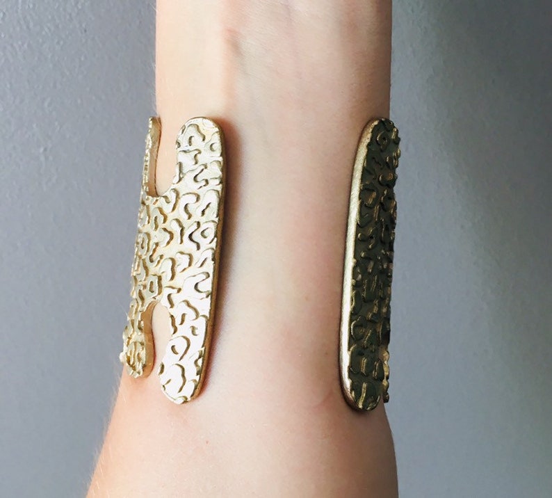 May include: Gold-toned metal cuff bracelet with a textured, abstract design. The bracelet is open and adjustable.