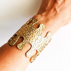 May include: A gold-toned metal cuff bracelet with a textured, abstract design. The bracelet has a wide, open design with a wavy, organic shape.