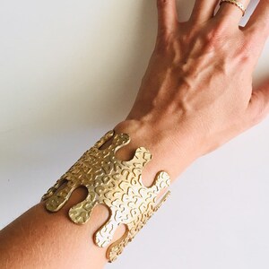 May include: A gold-toned metal bracelet with a unique, abstract design. The bracelet has a textured surface and a slightly open design.