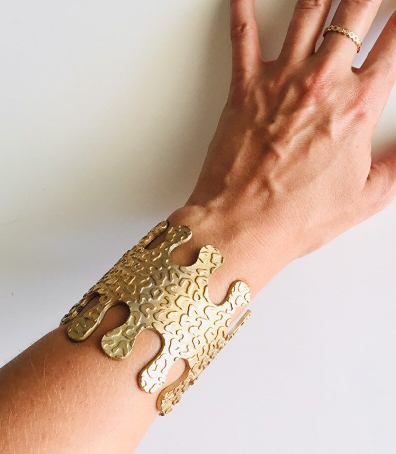May include: A gold-toned metal bracelet with a unique, abstract design. The bracelet has a textured surface and a slightly open design.