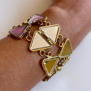 Peut inclure: Bracelet doré avec des segments triangulaires et miroités en rose, crème et jaune. Les segments sont reliés par de petits connecteurs circulaires. Le bracelet est porté au poignet.
