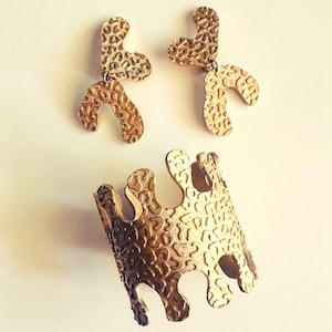 May include: A set of gold-toned jewelry featuring a pair of earrings and a bracelet. The earrings are shaped like abstract forms with a textured surface. The bracelet is a wide band with a similar textured surface and an irregular shape.