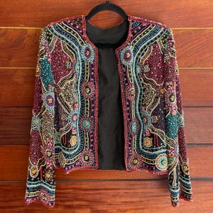 May include: A vibrant, open-front jacket adorned with detailed beadwork in pink, teal, gold, and black. The jacket has a black lining and is hung on a black hanger against a wooden backdrop. The design incorporates floral and geometric patterns.