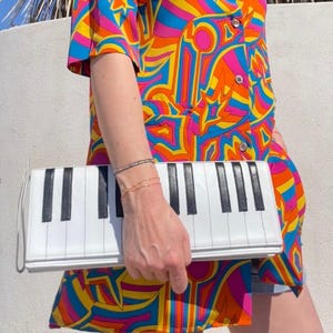 Peut inclure: Pochette blanche avec un motif de clavier de piano. La pochette a des touches noires et blanches et une petite poignée. La personne porte une chemise colorée à motifs avec des teintes orange, jaune, bleu et rose.