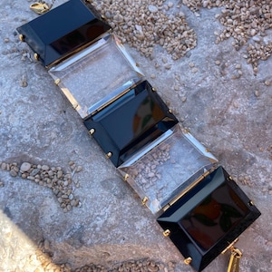 May include: A rectangular bracelet with alternating black and clear panels, accented with gold-colored hardware. The bracelet is made of a glossy material and has a clasp closure. The bracelet is lying on a stone surface.