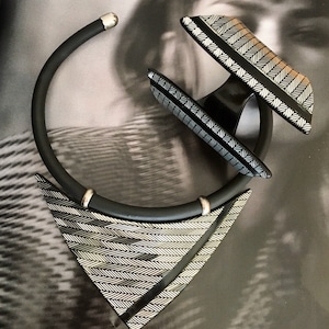 Può includere: Una collana geometrica nera e argentata con un ciondolo triangolare e un braccialetto abbinato. Il ciondolo e il braccialetto presentano un motivo a chevron.