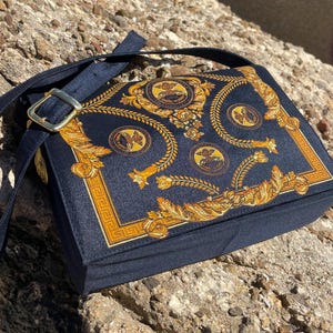 Puede incluir: Un bolso de hombro negro con un diseño estampado en oro y negro. El bolso tiene una correa larga y un cierre de solapa.